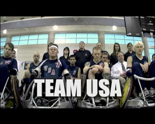 Front shot of four men on the US Paralympics team in quad rugby wheelchairs sitting ostensibly on the side of a court with supporters behind them. White text reads TEAM USA