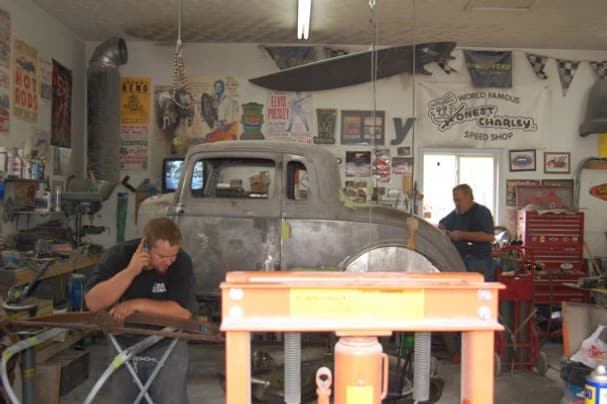 two men working in a crowded garage, rebuilding an antique car, with one on the phone