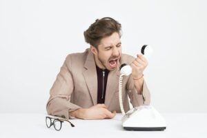White man in tan blazer sitting at whtie table with white background,  yelling into a white landline phone