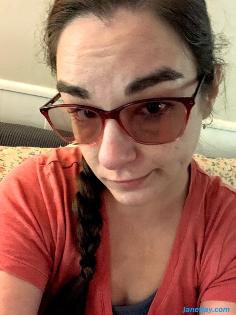 Picture of Janet Jay, a white girl with a pink shirt, a side braid and reddish sunglasses, looking at the camera with a look of "welp, this is it"