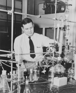 B&W, a man faces the camera 3/4, sitting at a desk and playing with scientific equipment. His right hand is missing, though this is not emphasized.