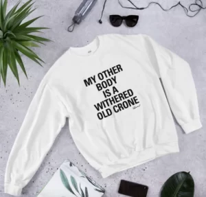 White sweatshirt lying on a grey background surrounded by plant, notebook, sunglasses etc, text on shirt reads "my other body is a withered old crone." Image from Reductress.com